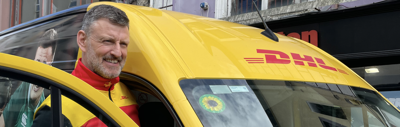 A male DHL delivery driver. His yellow DHL van is behind him. It has a Sunflower sticker on the windscreen 