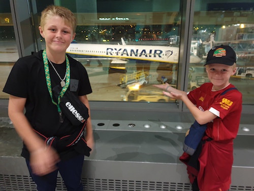 Two young white boys at the airport. They look happy. One wears a Sunflower lanyard and there is an aeroplane on the tarmac visible through the window. 