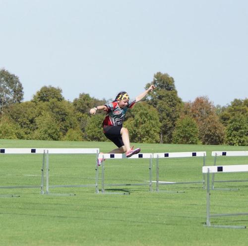 Lily, a young white girl, competing in a hurdles race on grass. She is mid air going over a hurdle. There are trees in the background. 