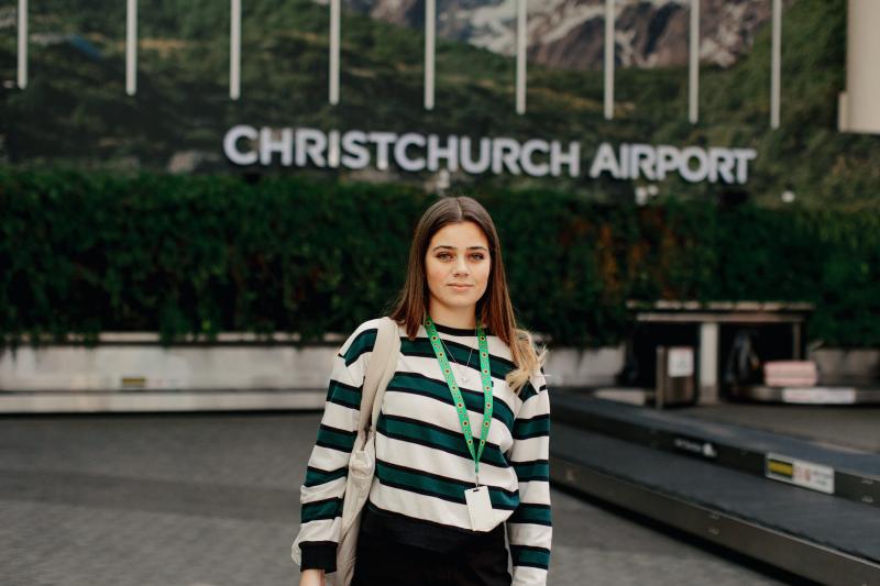 A young, white female with long dark hair stands in front of Christchurch airport. She wears a green and white striped jumper and a Sunflower lanyard