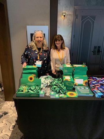 Ruth and Lynn at the Airport Customer Experience Symposium. There are Sunflower items on display on top of a table in front of the ladies. 