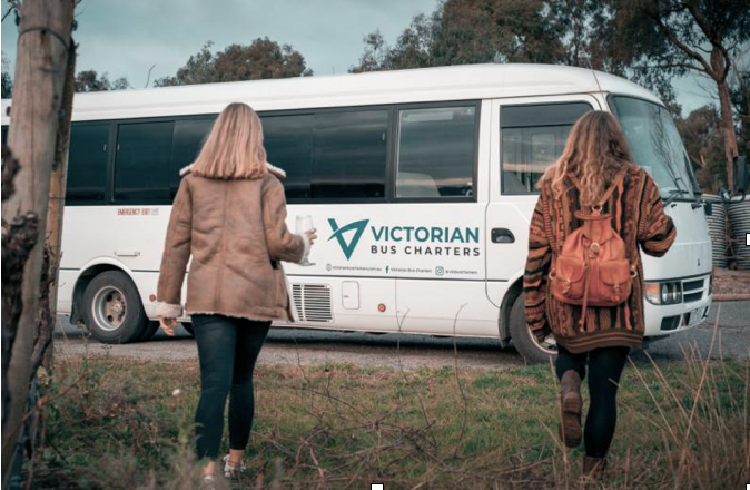 Two white women with their backs to us walk towards a white Victorian Bus charters coach. They wear brown coats and one wears a brown backpack
